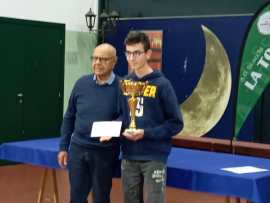 Two individuals standing together in a room, one holding a trophy and the other a certificate. The background features a moon graphic and some banners.