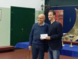 Two men standing together in a room, one is receiving a certificate or award from the other. They are smiling, with a table in the background displaying trophies and a blue cloth.