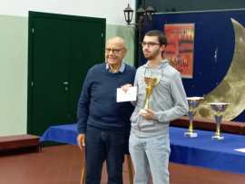 Two men standing together at an awards ceremony, one holding a trophy and a check, with a blue table and trophies in the background.