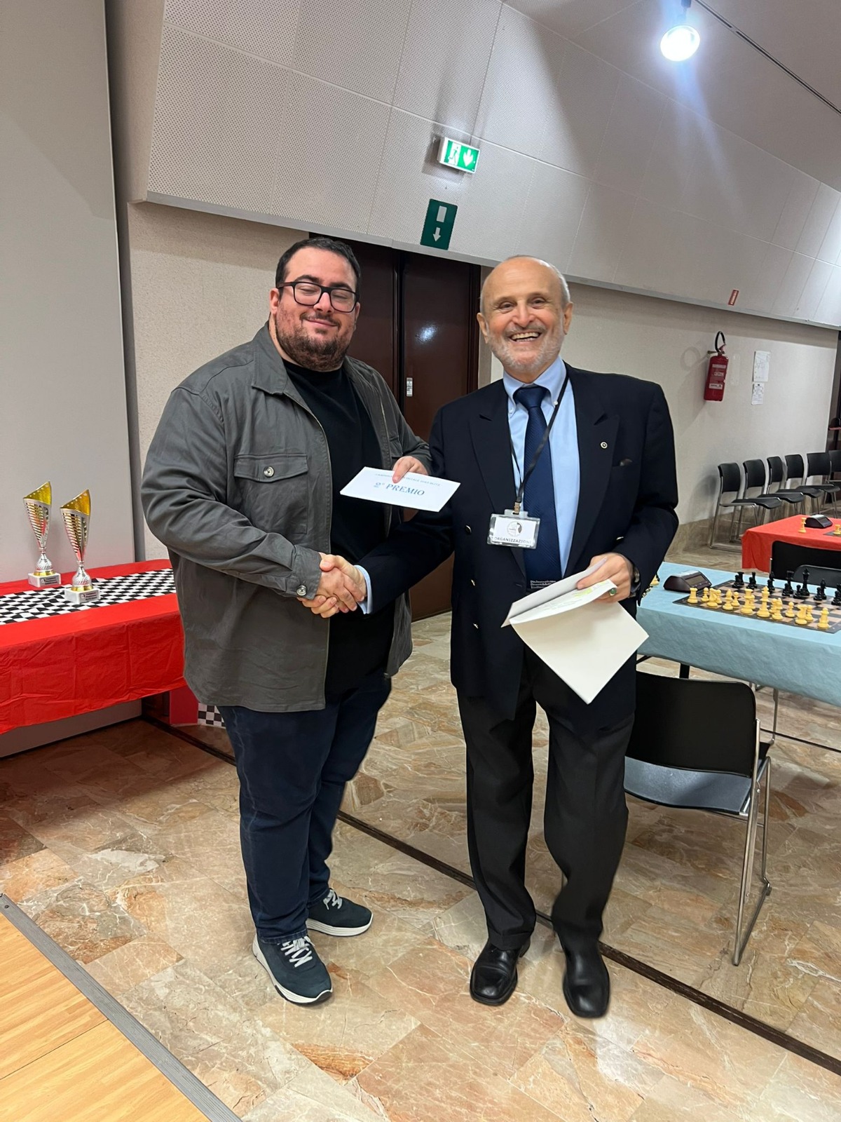 Due uomini si stringono la mano in un evento di premiazione, con uno che riceve un documento o un premio. Sullo sfondo, tavoli con attrezzature scacchistiche e decorazioni.