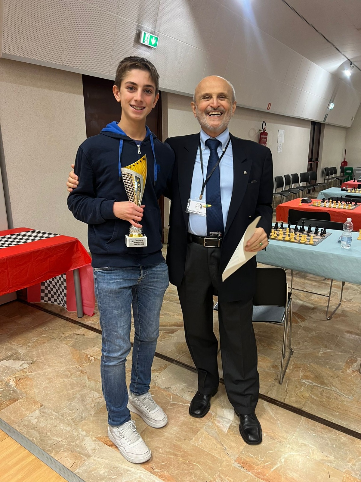 Due uomini sorridenti posano per una foto, uno è un giovane con un trofeo in mano e l'altro è un uomo più anziano con un foglio. Sullo sfondo si intravedono tavoli con materiali di scacchi.