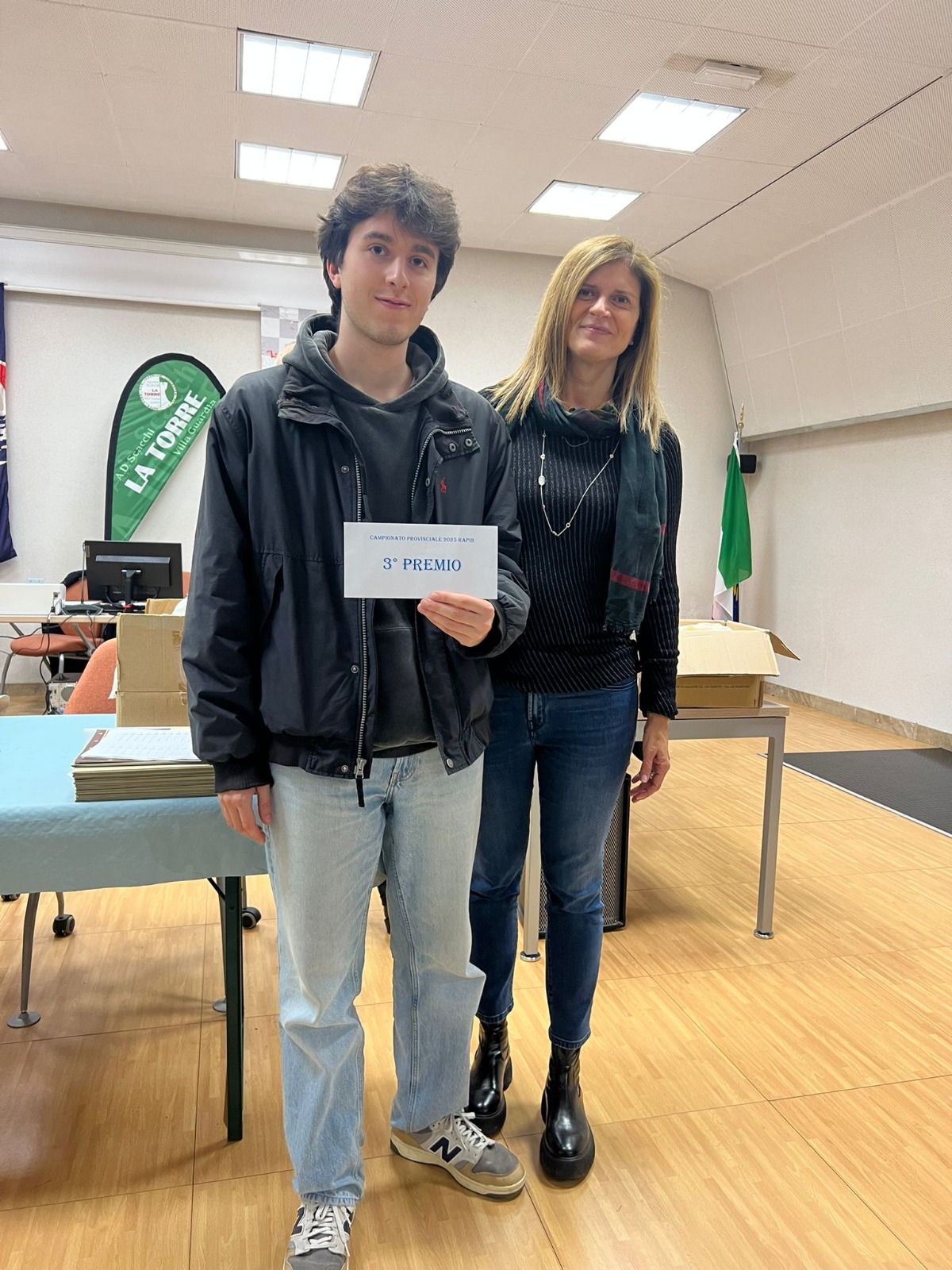 Un giovane campione di scacchi premiato in un evento con un premio in mano, affiancato da una donna in un ambiente di competizione.