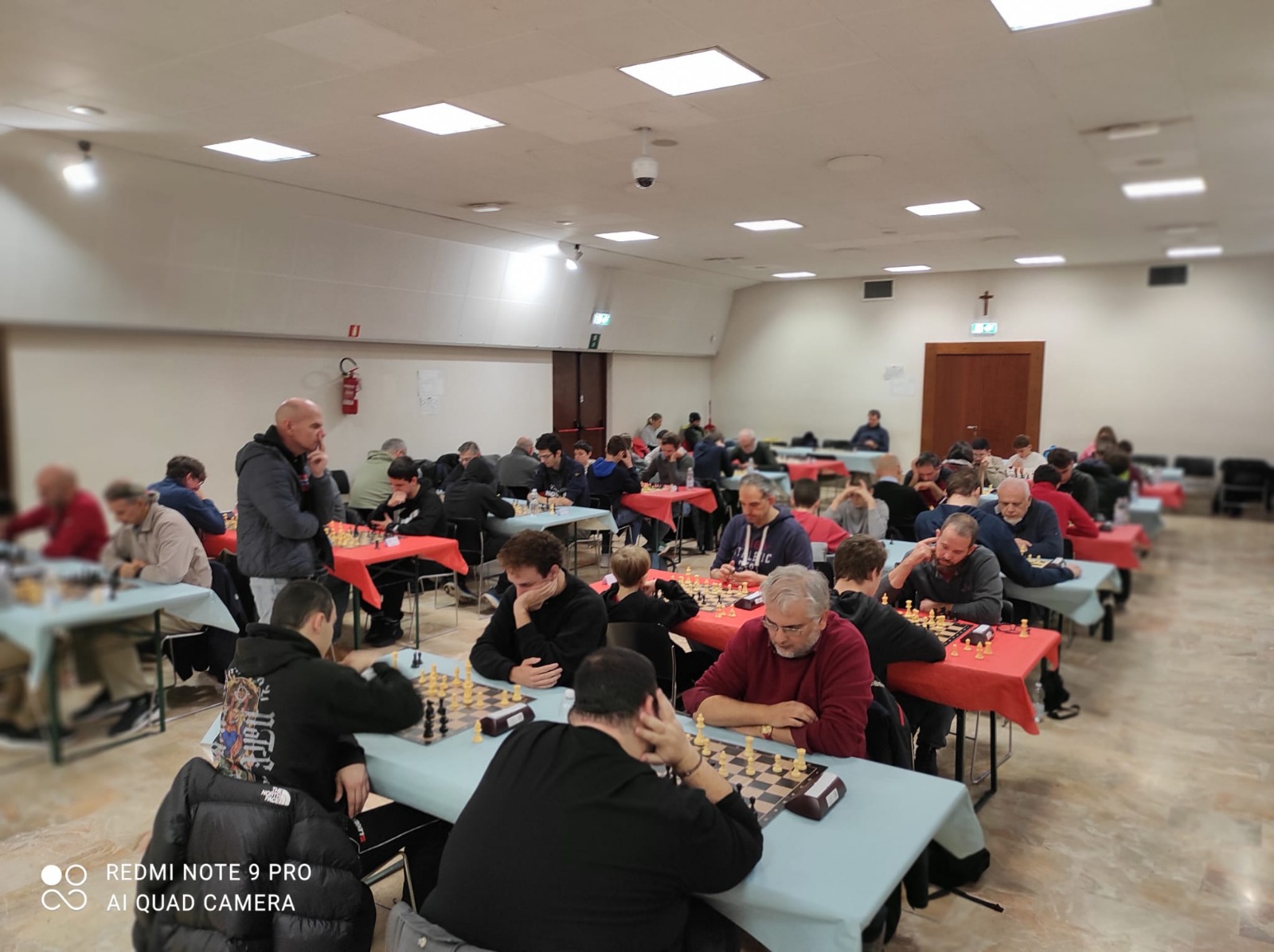 A crowded chess tournament taking place in a well-lit hall with players deeply focused on their games. Tables are set with chess boards and pieces, and participants show varying expressions of concentration.