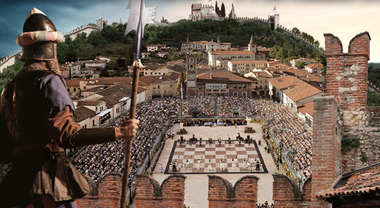 Un soldato in armatura osserva una grande piazza affollata dove si svolge un torneo di scacchi all'aperto, con edifici storici sullo sfondo.