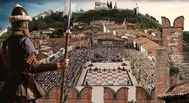 Un soldato in armatura osserva una grande piazza affollata dove si svolge un torneo di scacchi all'aperto, con edifici storici sullo sfondo.