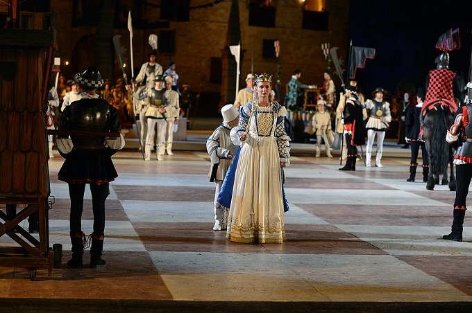 Rappresentazione storica con un gruppo di persone in costume d'epoca, tra cui una donna in abito bianco e un bambino, sullo sfondo di un campo con cavalieri e soldati.