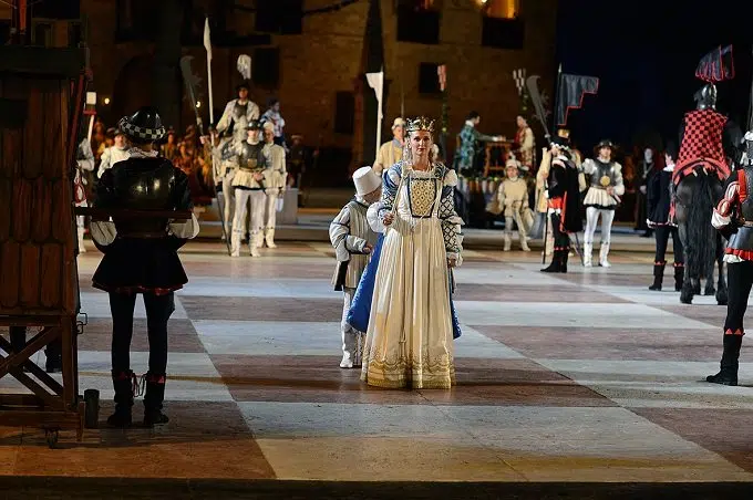 Rappresentazione storica con un gruppo di persone in costume d'epoca, tra cui una donna in abito bianco e un bambino, sullo sfondo di un campo con cavalieri e soldati.