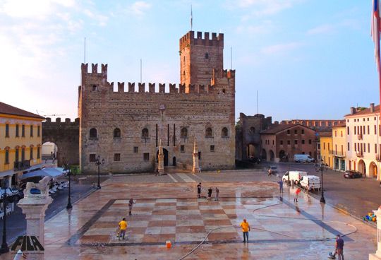 Vista del castello medievale in piazza con persone e veicoli.
