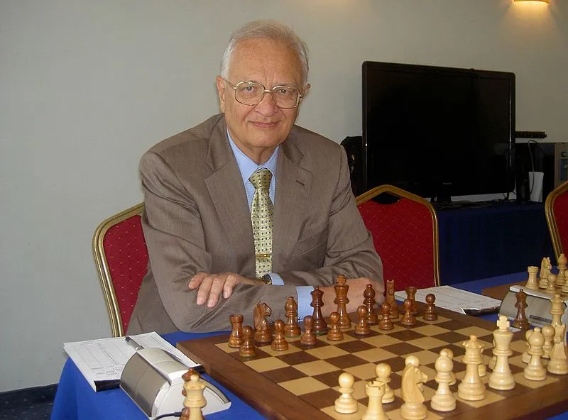 Un uomo anziano seduto a un tavolo da scacchi con pezzi di scacchi bianchi e neri, indossa un abito elegante e sorride.