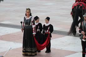 Due bambini in abiti tradizionali portano un drappo rosso, mentre una donna in abito folkloristico li accompagna.