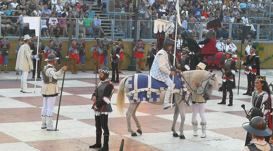 Un torneo medieval con cavalieri, soldati e un pubblico numeroso, in un'arena colorata.