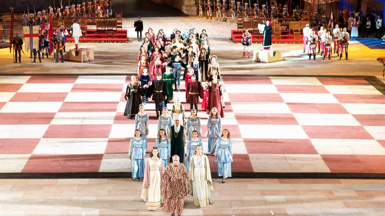 Un grande spettacolo con diversi performer in costume che sfilano su un grande palcoscenico con un pavimento a scacchiera.