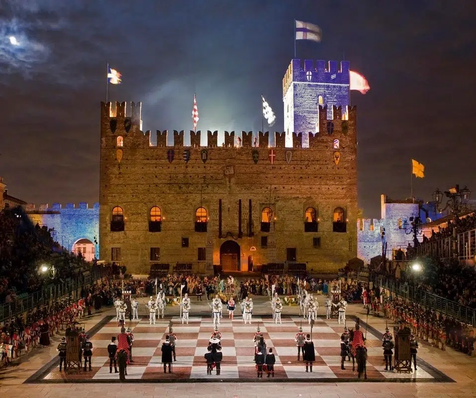 Una grande scacchiera allestita davanti a un castello illuminato durante la notte, con figuranti in costumi storici e spettatori in attesa.