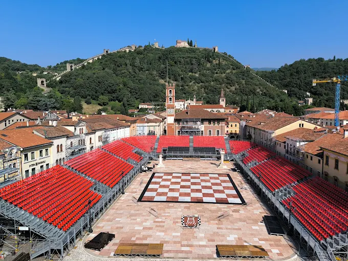 Vista panoramica di una piazza con tribune rosse e un grande scacchiere, circondata da edifici storici e colline verdi.