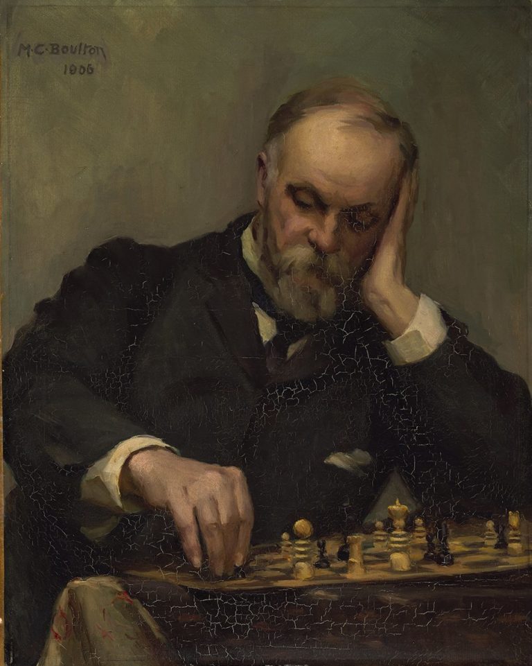 Un uomo anziano, con la barba e i capelli grigi, sta pensando mentre gioca a scacchi. Indossa un completo nero e ha un'espressione concentrata.