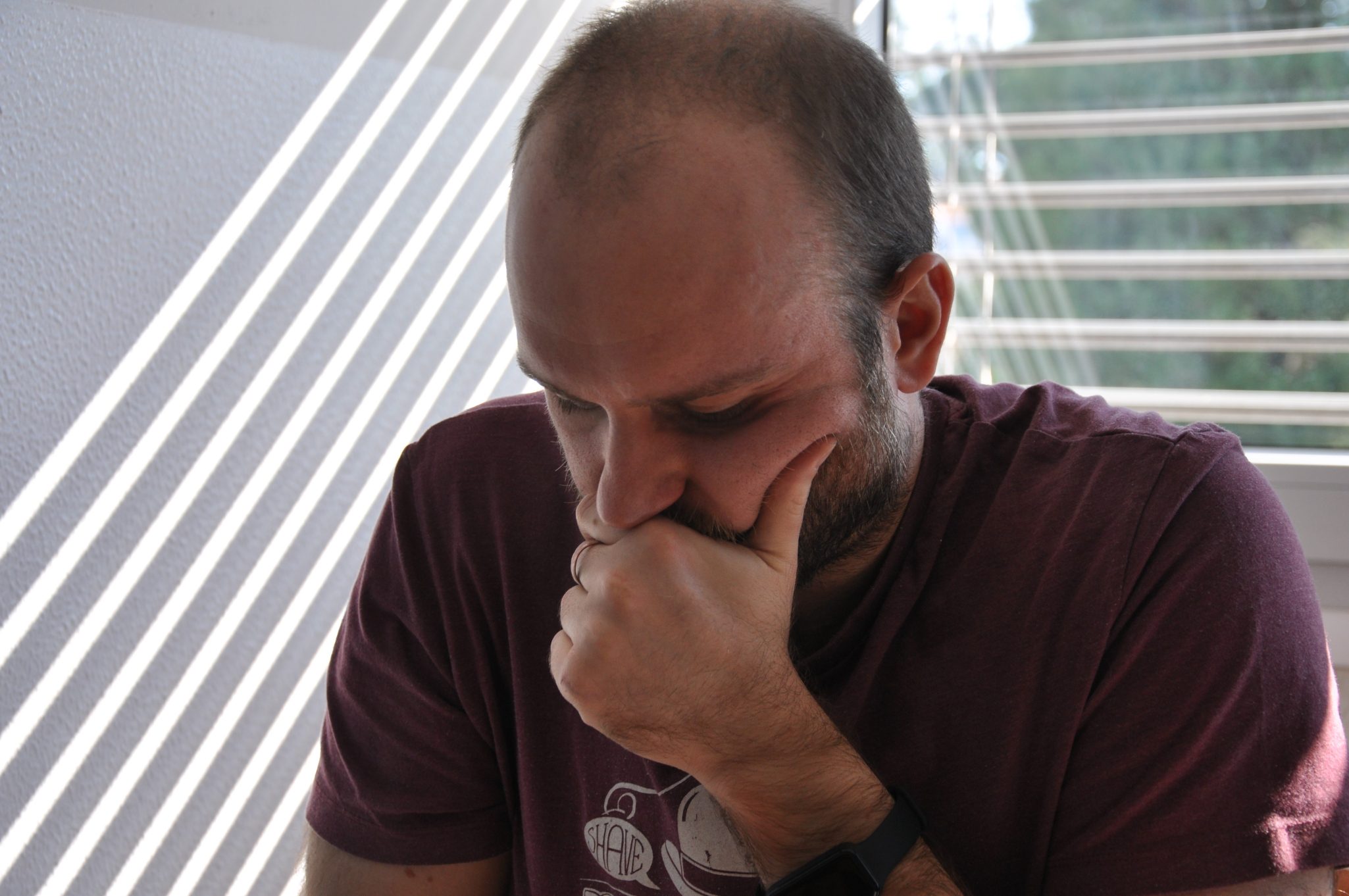 Uomo pensieroso con mano sulla bocca, illuminato da luce naturale che entra da una finestra.