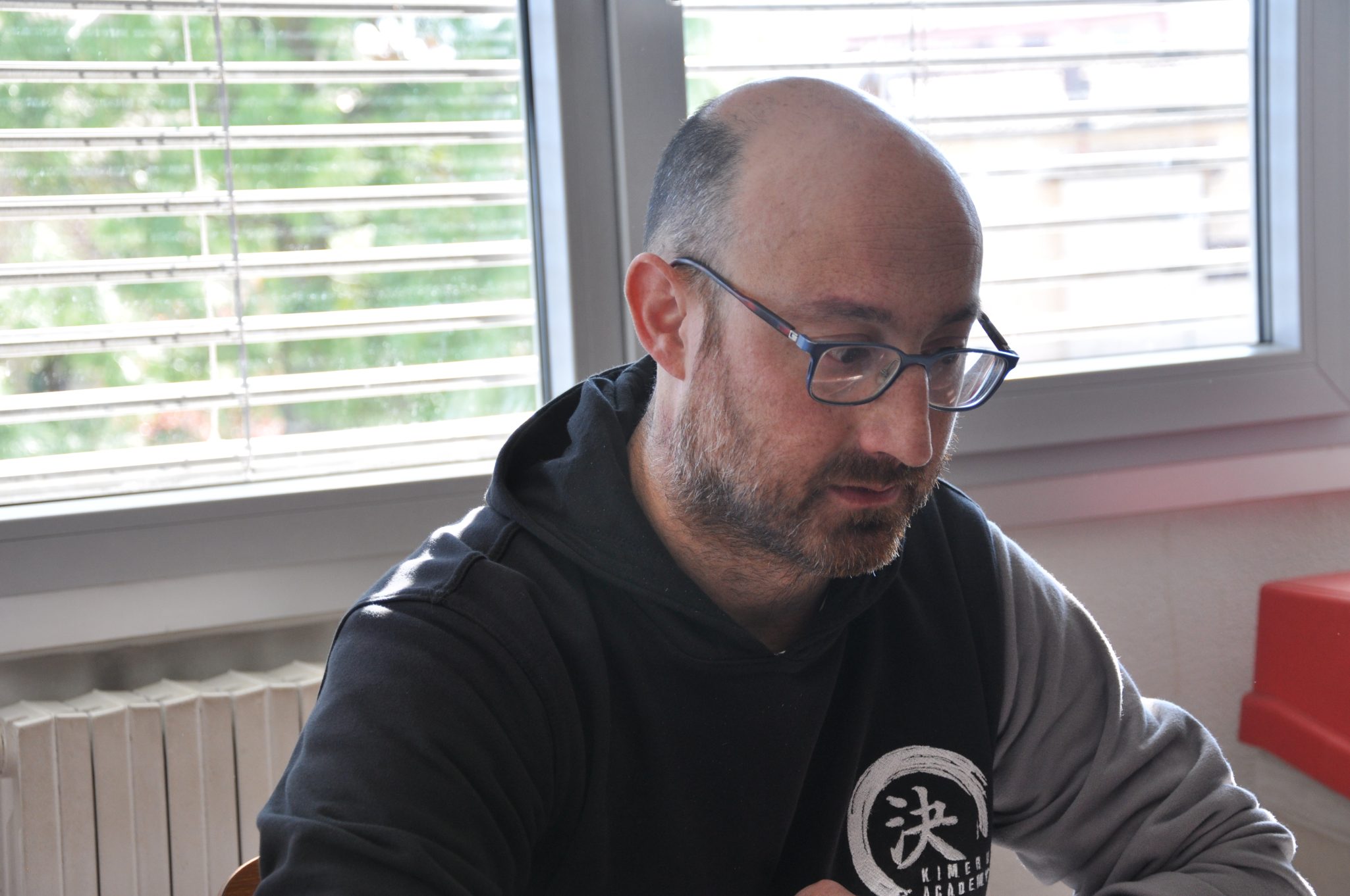 Un uomo calvo con occhiali, seduto e concentrato, indossa una felpa nera con dettagli grigi. Sullo sfondo si intravede una finestra con persiane e un termosifone.