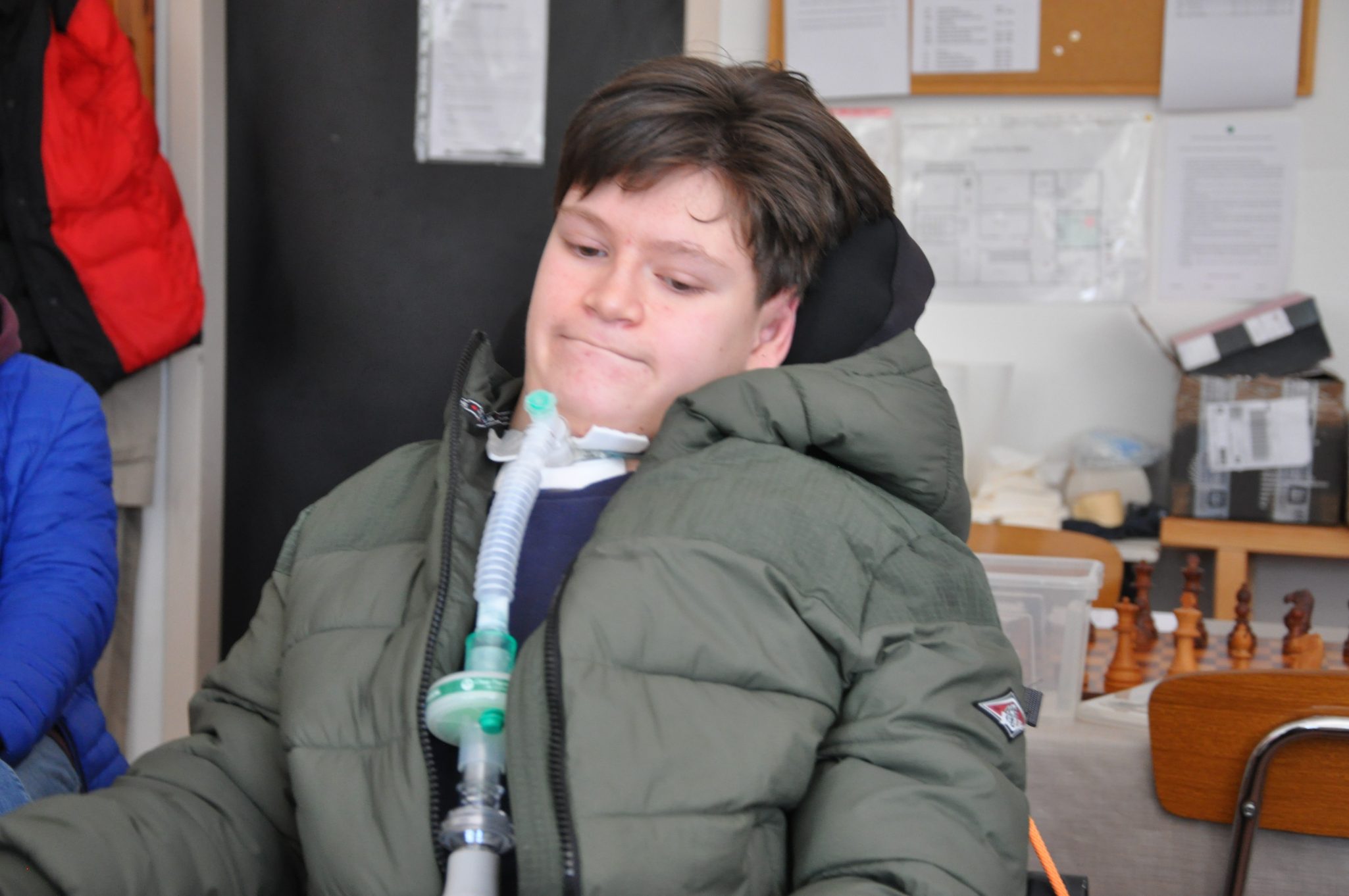 Ragazzo seduto in una stanza, indossando un giubbotto pesante. Ha un dispositivo di ventilazione collegato e appare pensieroso.