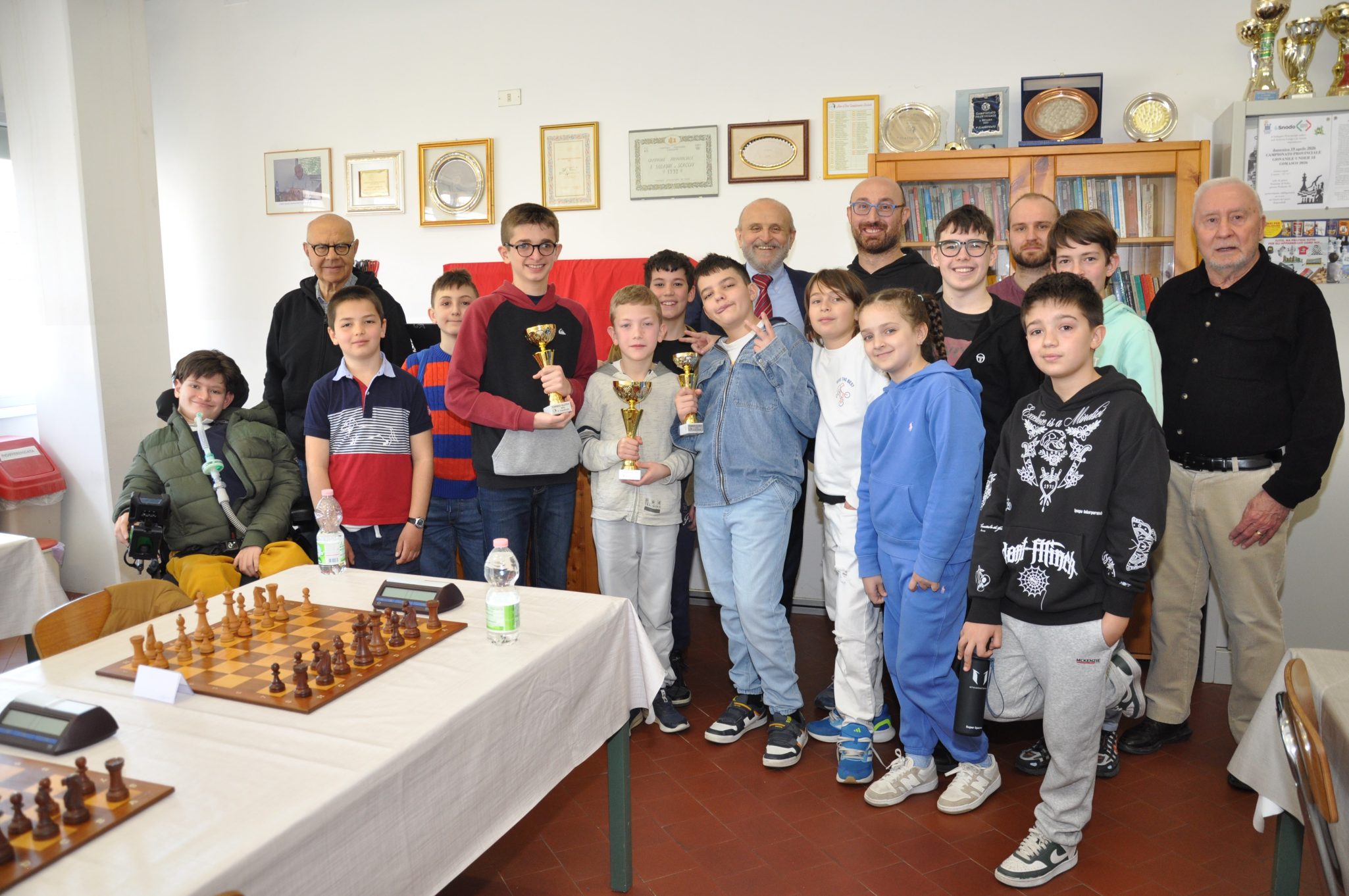 Gruppo di giovani e adulti sorridenti dopo un torneo di scacchi, con trofei in mano. Tavoli con scacchiere e trofei sullo sfondo.