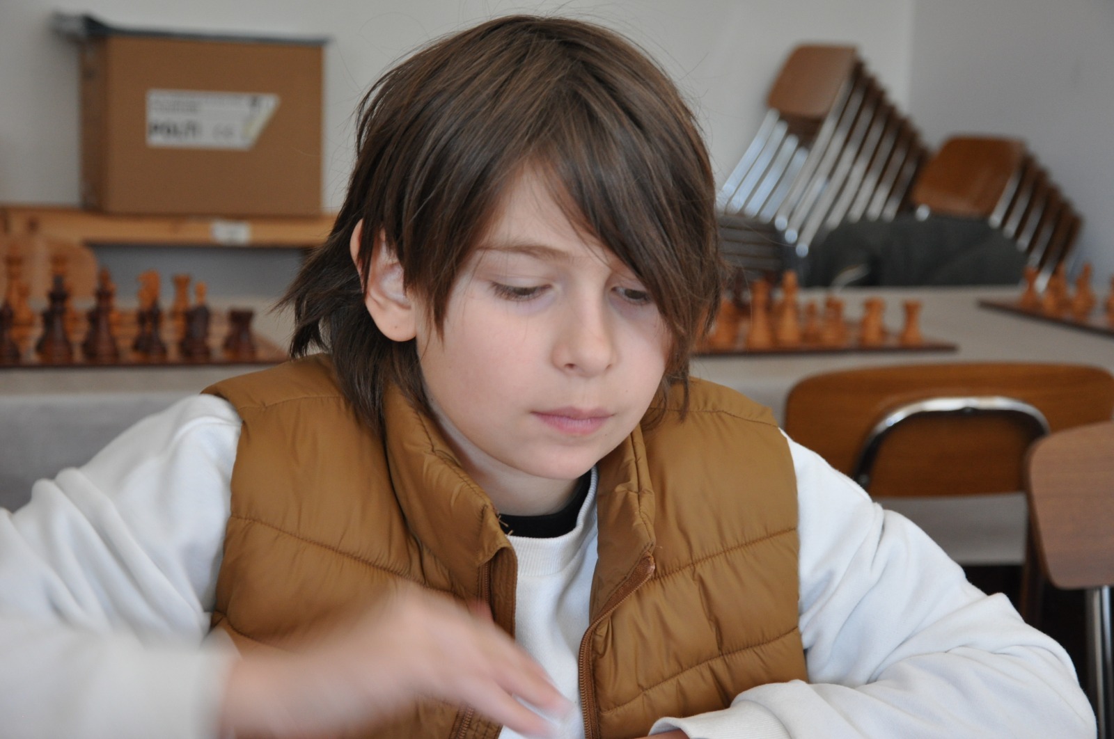 Ragazzo con capelli castani, vestito con una giacca marrone, mentre concentra l'attenzione su un'attività, con una scacchiera visibile sullo sfondo.