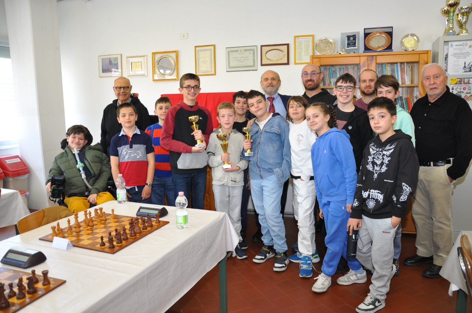 Gruppo di giovani giocatori di scacchi sorridenti con trofei in mano, in una sala decorata con premi e certificati.