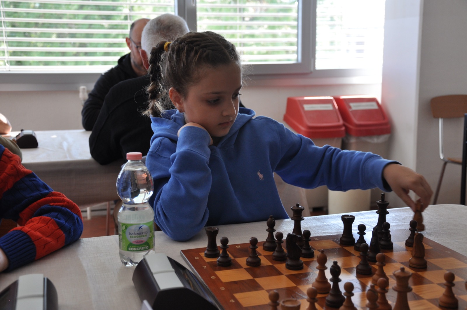 Una bambina gioca a scacchi in un ambiente tranquillo, mentre altri giocatori sono seduti in background.