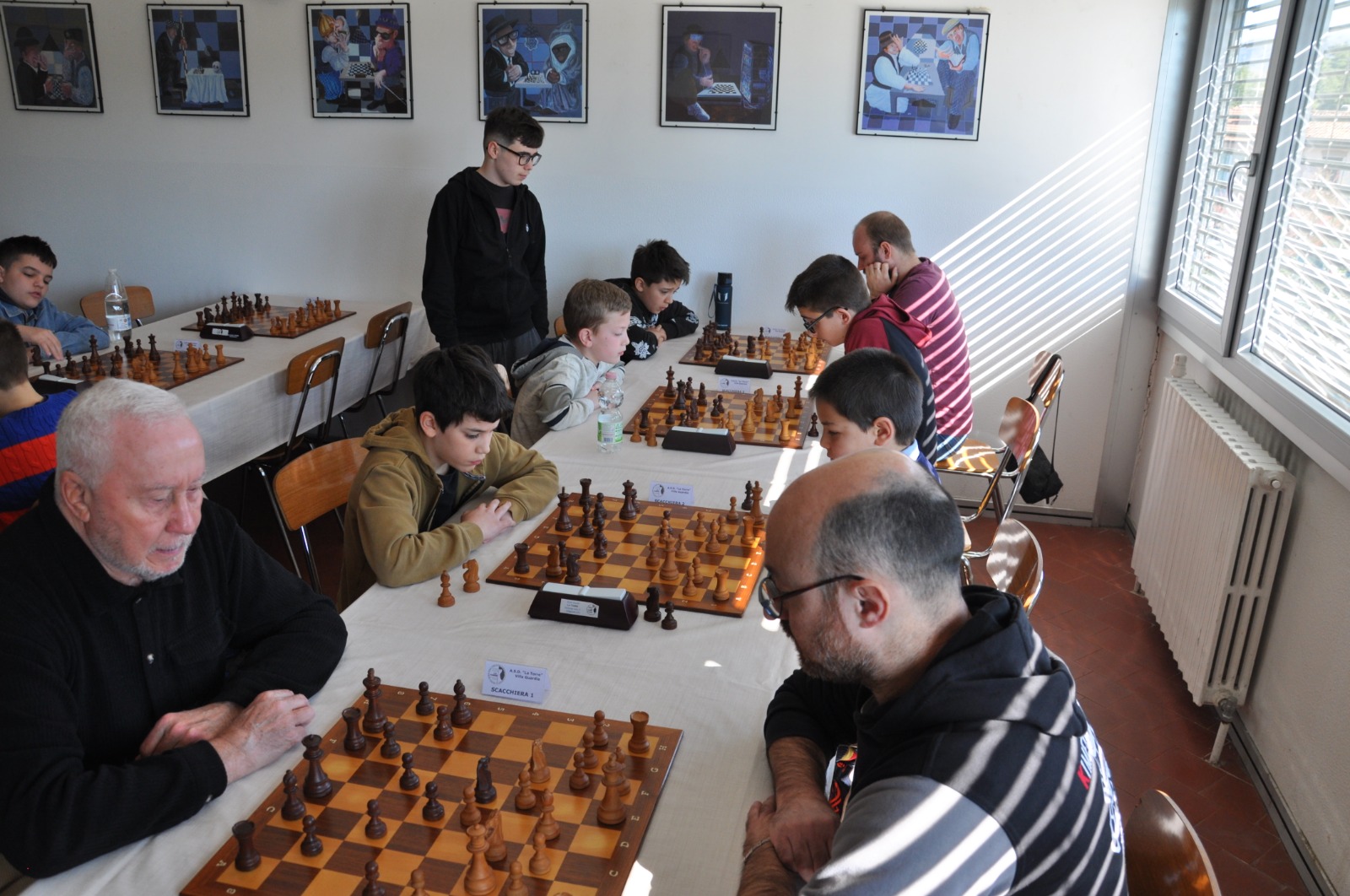 Un torneo di scacchi con giocatori di diverse età seduti a tavoli, concentrati sulle loro partite.