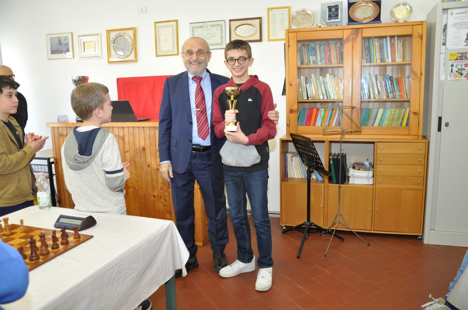 Un ragazzo con un trofeo sorride accanto a un uomo anziano in una stanza con libri e riconoscimenti, mentre altri giovani lo osservano.