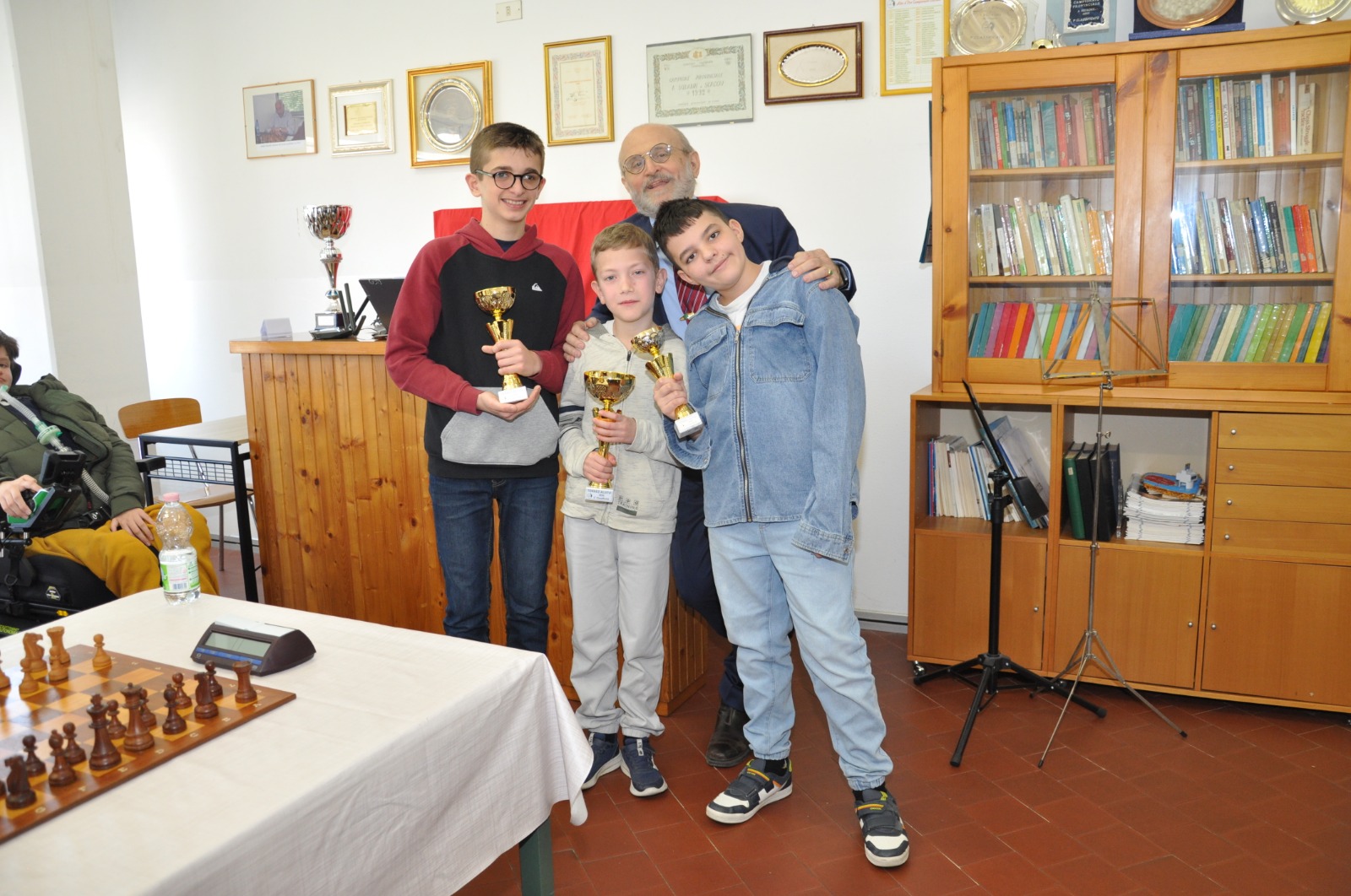 Tre ragazzi sorridenti posano con trofei dopo un torneo di scacchi, con un adulto che si trova dietro di loro. Sullo sfondo, una libreria con riconoscimenti.