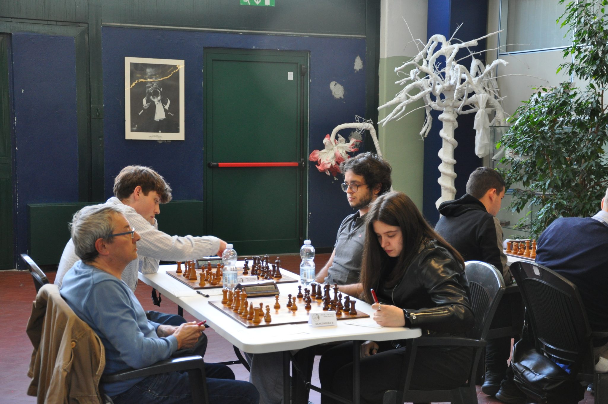 Un torneo di scacchi con diversi partecipanti seduti a tavoli, mentre alcuni giocatori stanno facendo le loro mosse e altri prendono appunti. Sullo sfondo si vede un albero decorativo e una foto appesa al muro.