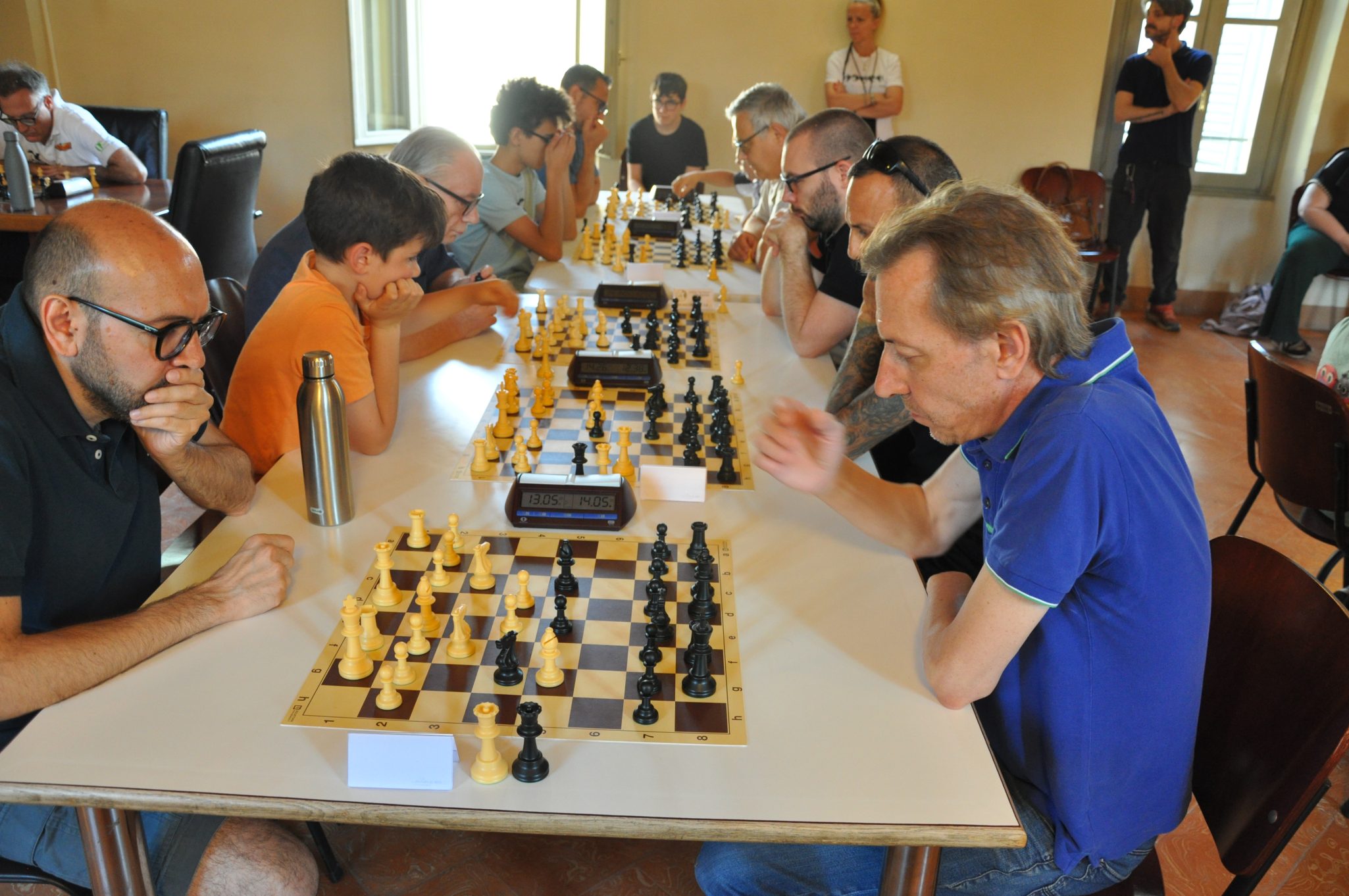 Un gruppo di persone gioca a scacchi in una stanza luminosa, con diverse scacchiere e cronometri da gioco sui tavoli.
