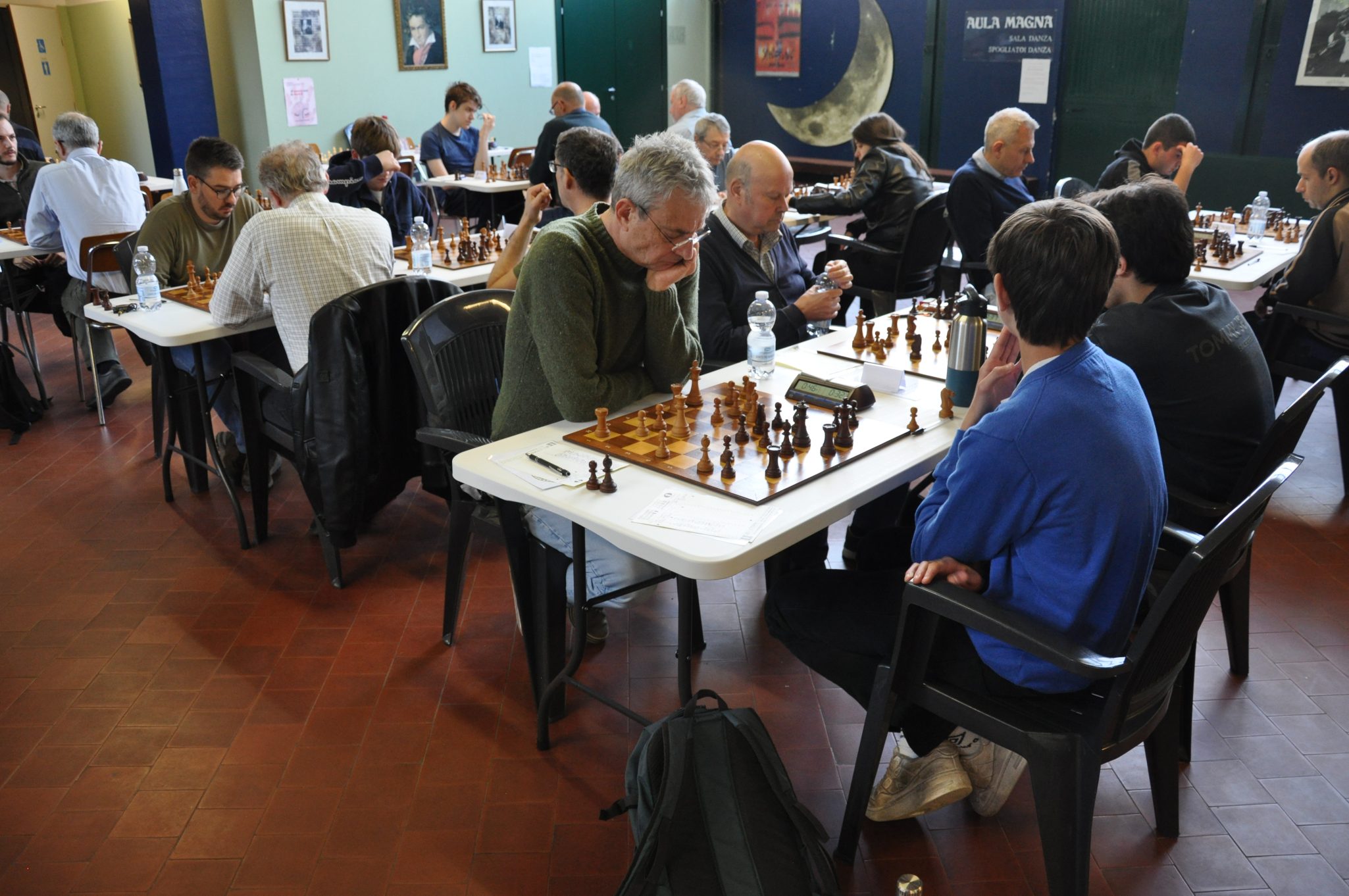 Un torneo di scacchi con partecipanti di diverse età seduti a tavoli, concentrati sulle loro partite in un ambiente indoor.