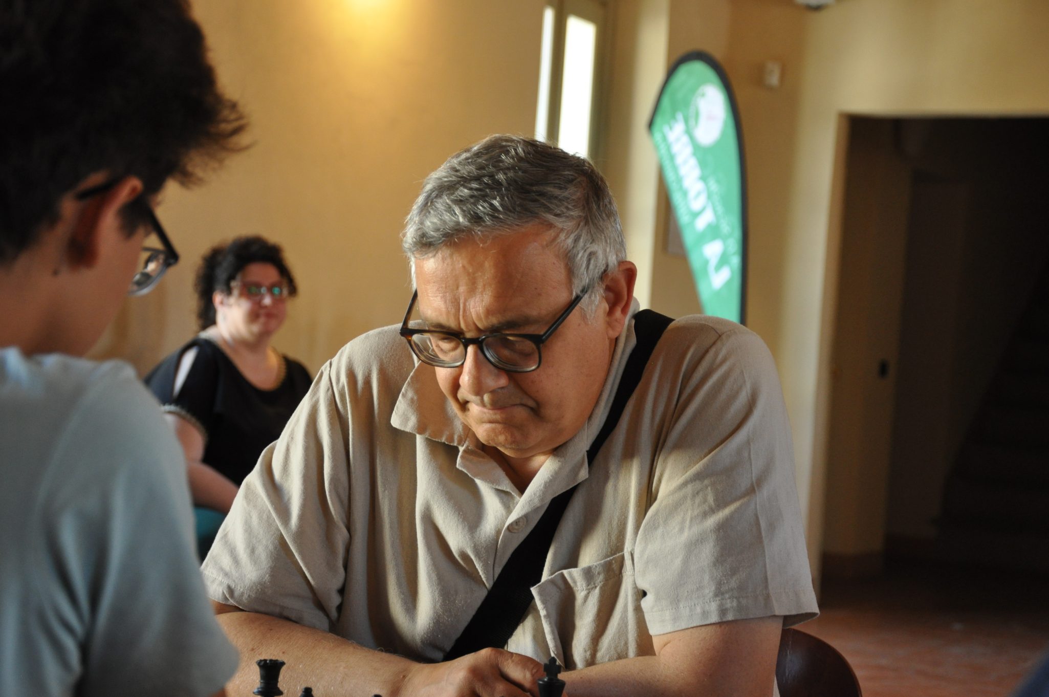 Due persone giocano a scacchi in un ambiente luminoso, con un uomo anziano concentrato sulla scacchiera e una donna che osserva sullo sfondo.