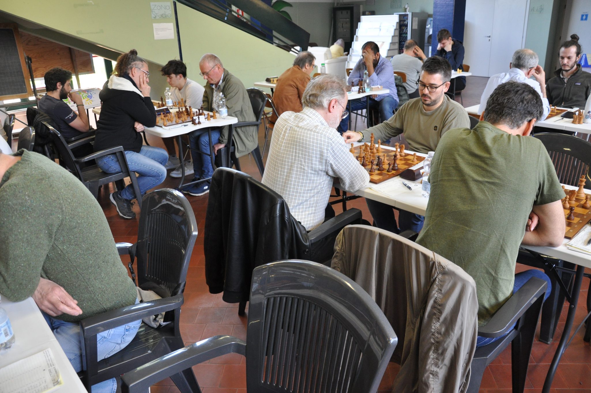 Giocatori di scacchi concentrati durante un torneo, seduti attorno a tavoli con scacchiere, in un ambiente pubblico.