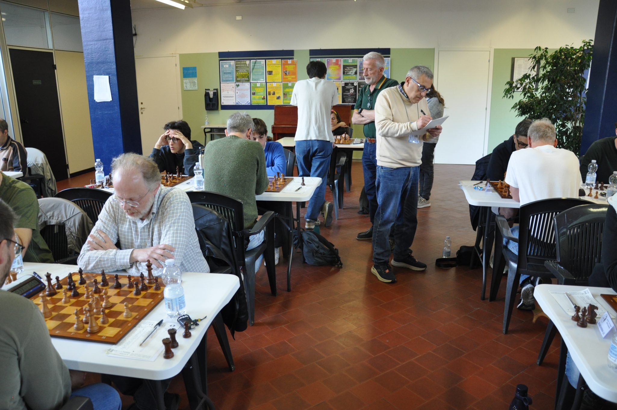 Una stanza con diverse persone che giocano a scacchi a tavoli. Alcuni giocatori sono concentrati sulla scacchiera, mentre altri stanno osservando o prendendo appunti. Sullo sfondo si vedono poster e una pianta.