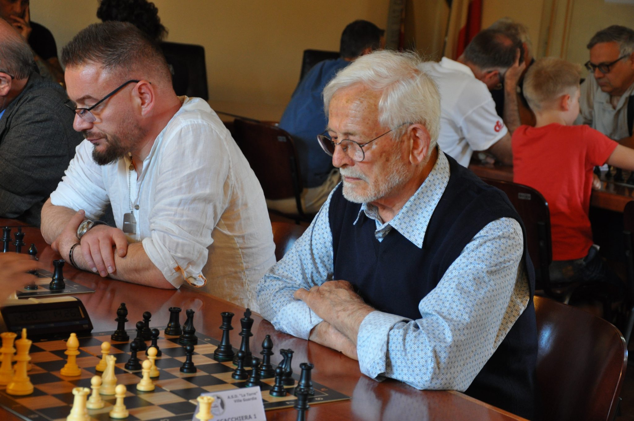 Due uomini stanno giocando a scacchi in un torneo. A sinistra, un uomo con barba e occhiali, vestito con una camicia bianca. A destra, un uomo anziano con capelli bianchi e occhiali, in attesa della sua mossa, con braccia incrociate.