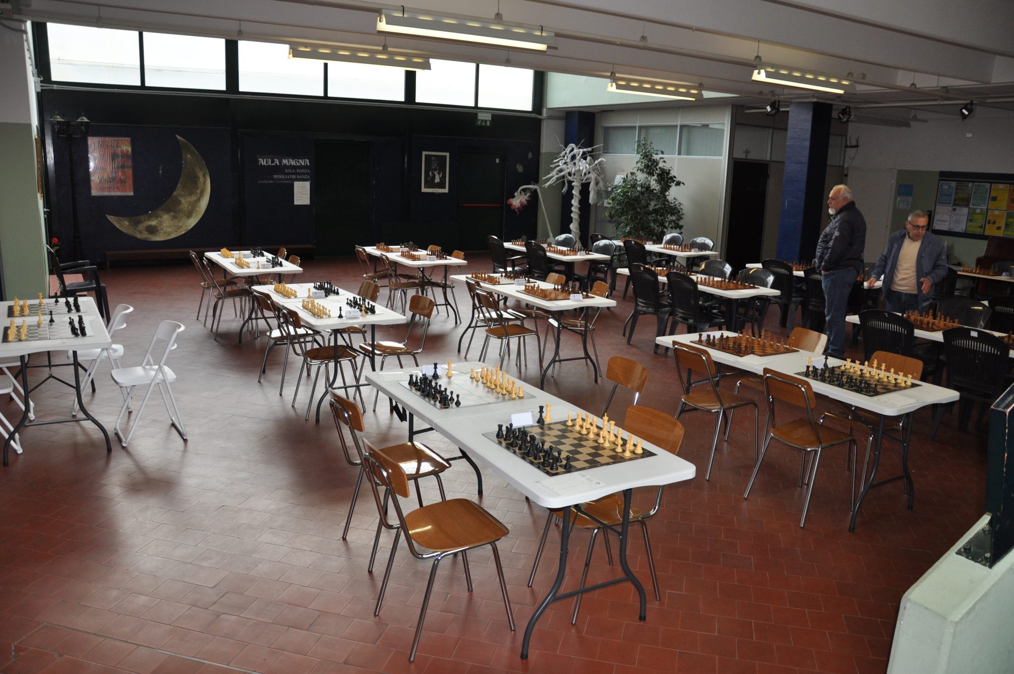 Una sala di gioco con tavoli disposti per il gioco degli scacchi, con pezzi neri e gialli pronti per le partite. Due uomini stanno esaminando i tavoli. Sullo sfondo si vede un grande muro decorato e piante in vaso.