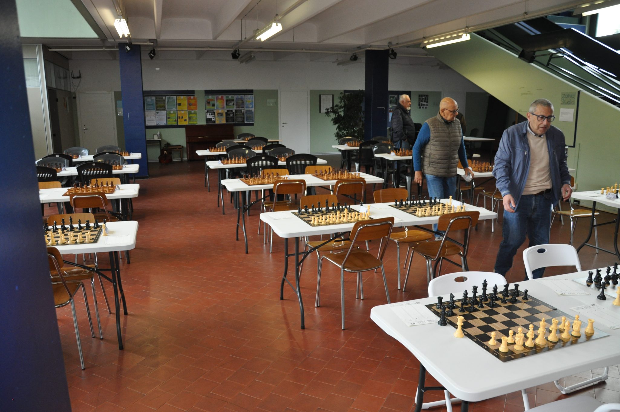 Una sala di gioco con tavoli e scacchiere pronte per una partita a scacchi, con alcuni giocatori che camminano tra i tavoli.