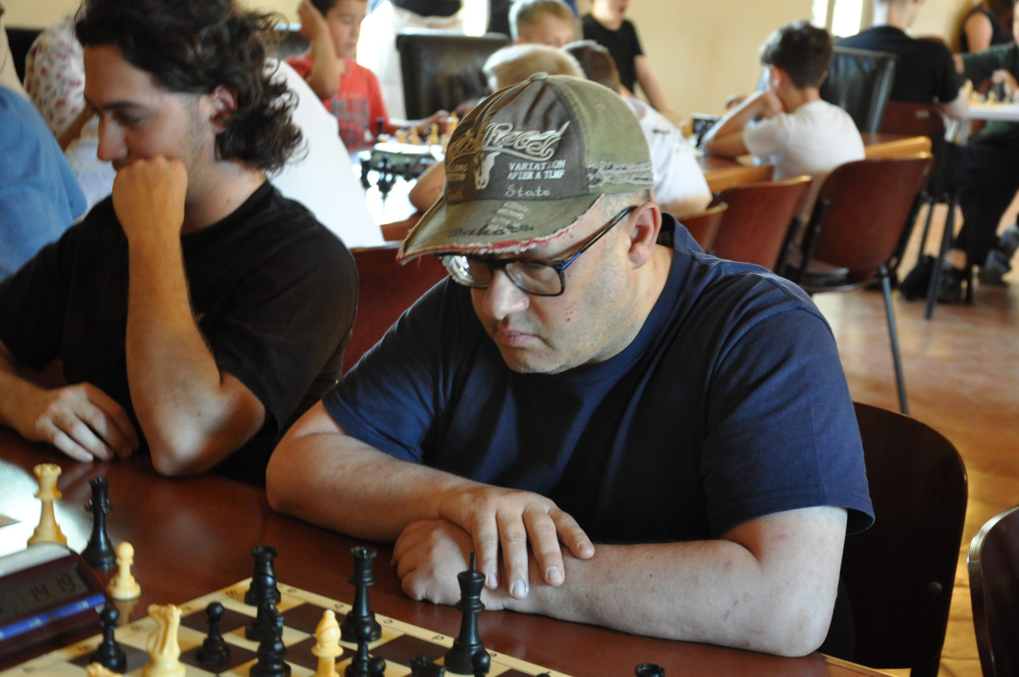 Un uomo con occhiali e un cappellino osserva una scacchiera durante una partita di scacchi in un ambiente affollato.
