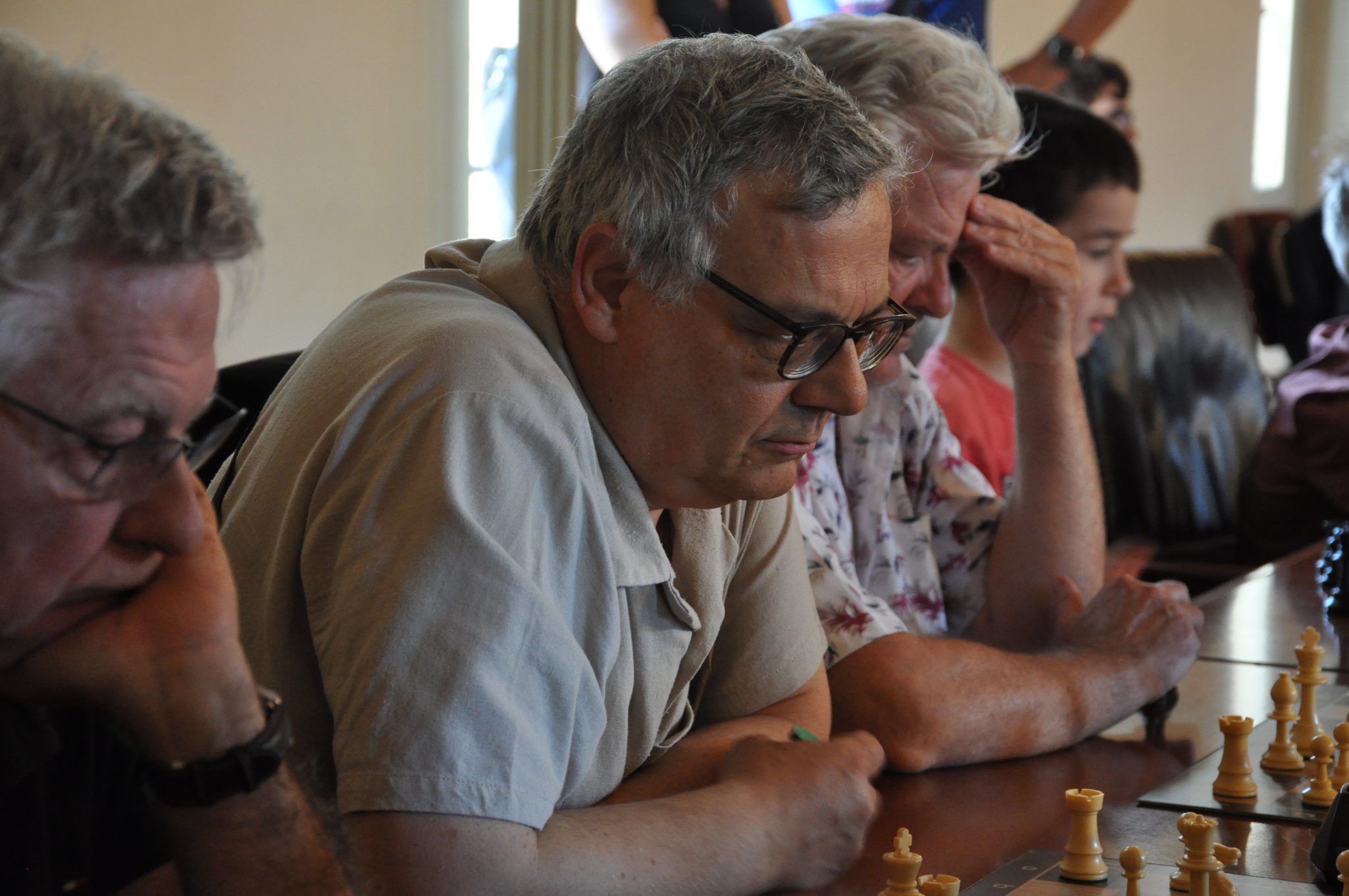 Un gruppo di persone gioca a scacchi, concentrati sulla partita. Si può vedere un uomo in primo piano che osserva una scacchiera con pezzi in legno.