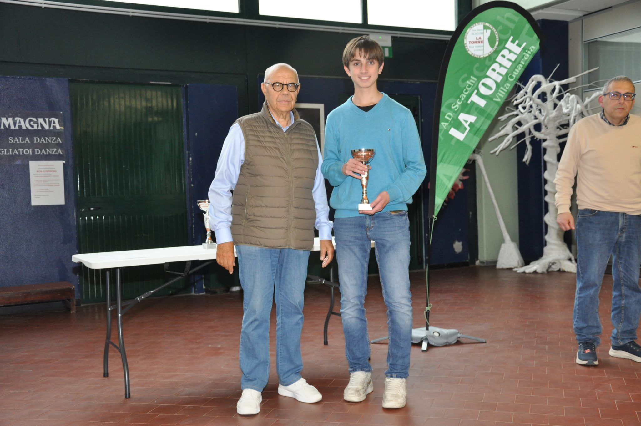 Un giovane premio vincitore tiene un trofeo, in compagnia di un adulto, in un'area di danza con una bandiera verde sullo sfondo.