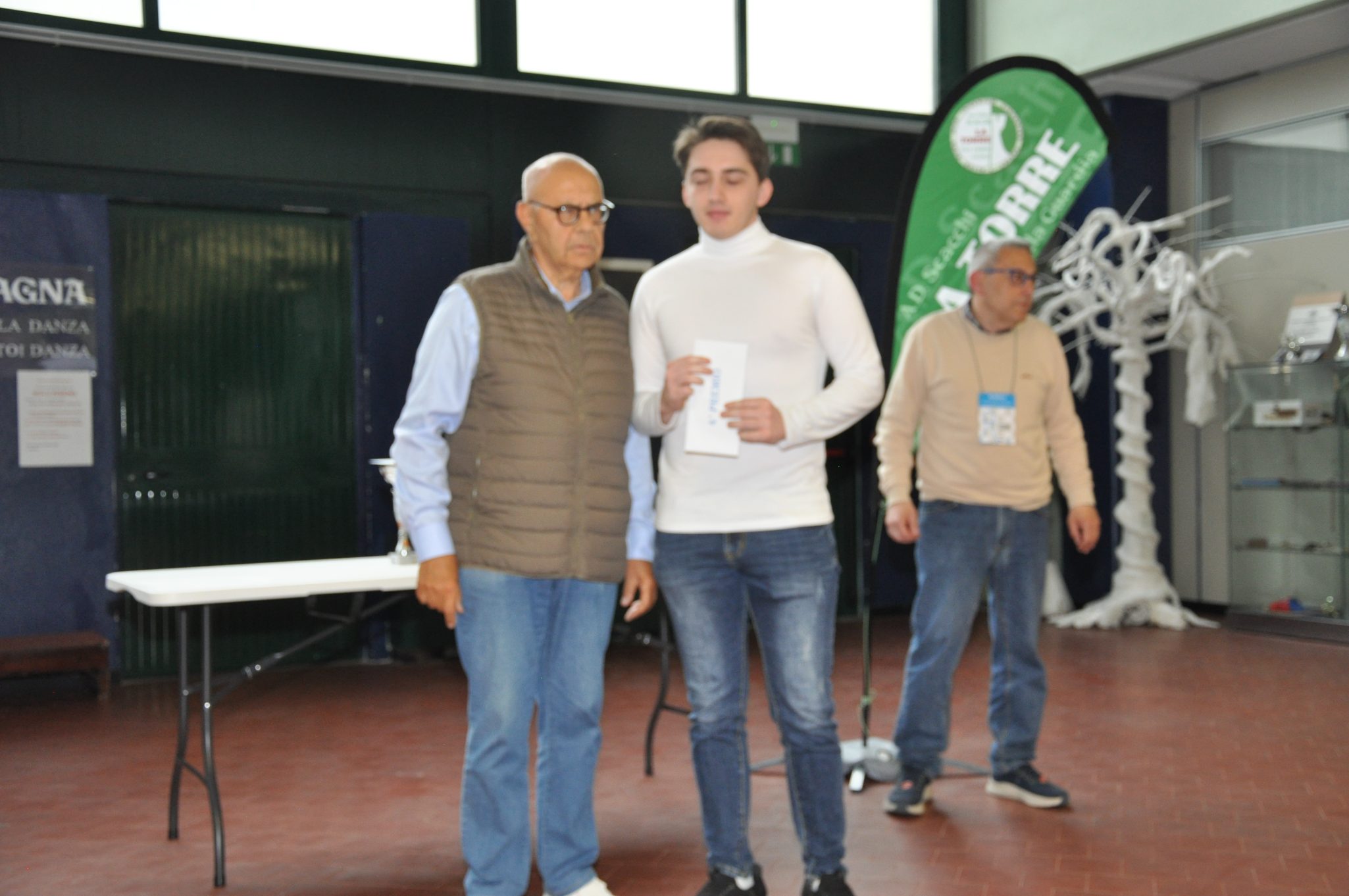 Due uomini in un evento, uno è in piedi con una busta di carta mentre l'altro lo guarda. Nell'ambiente ci sono tavoli e decorazioni.