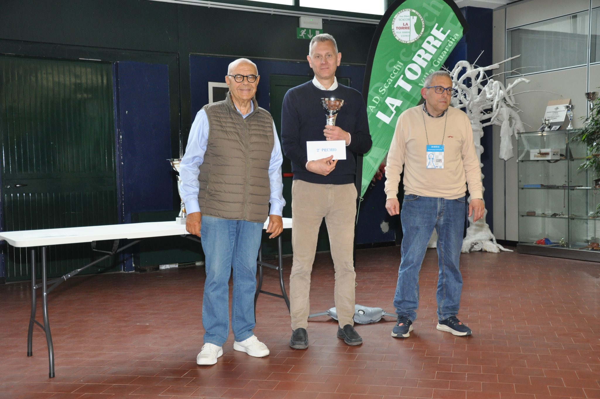 Tre uomini che partecipano a una cerimonia di premiazione, con un trofeo e un cartello che indica il premio.