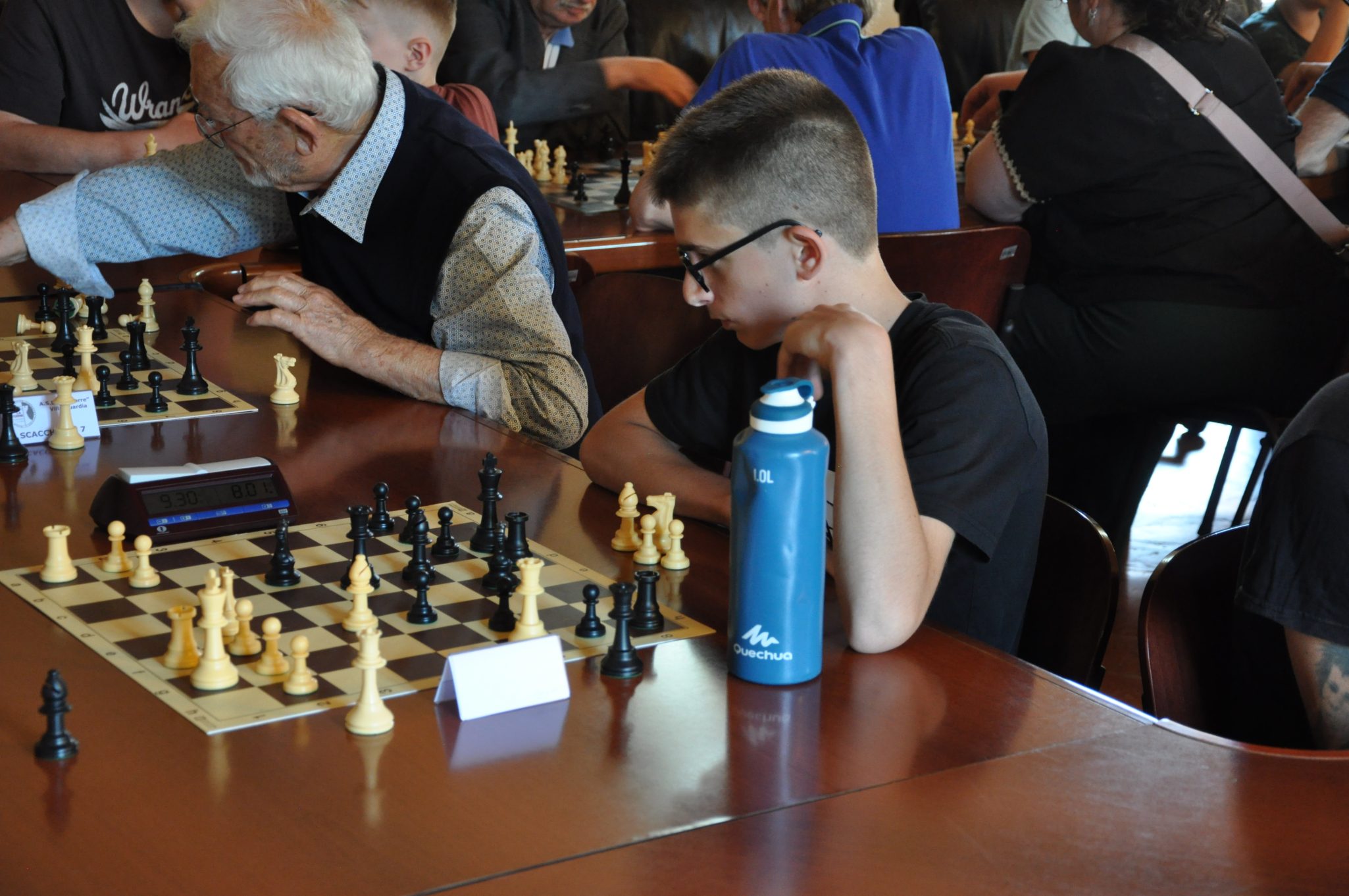 Due giocatori di scacchi in un torneo, un anziano e un ragazzo, con tavoli e scacchiere in un ambiente affollato.