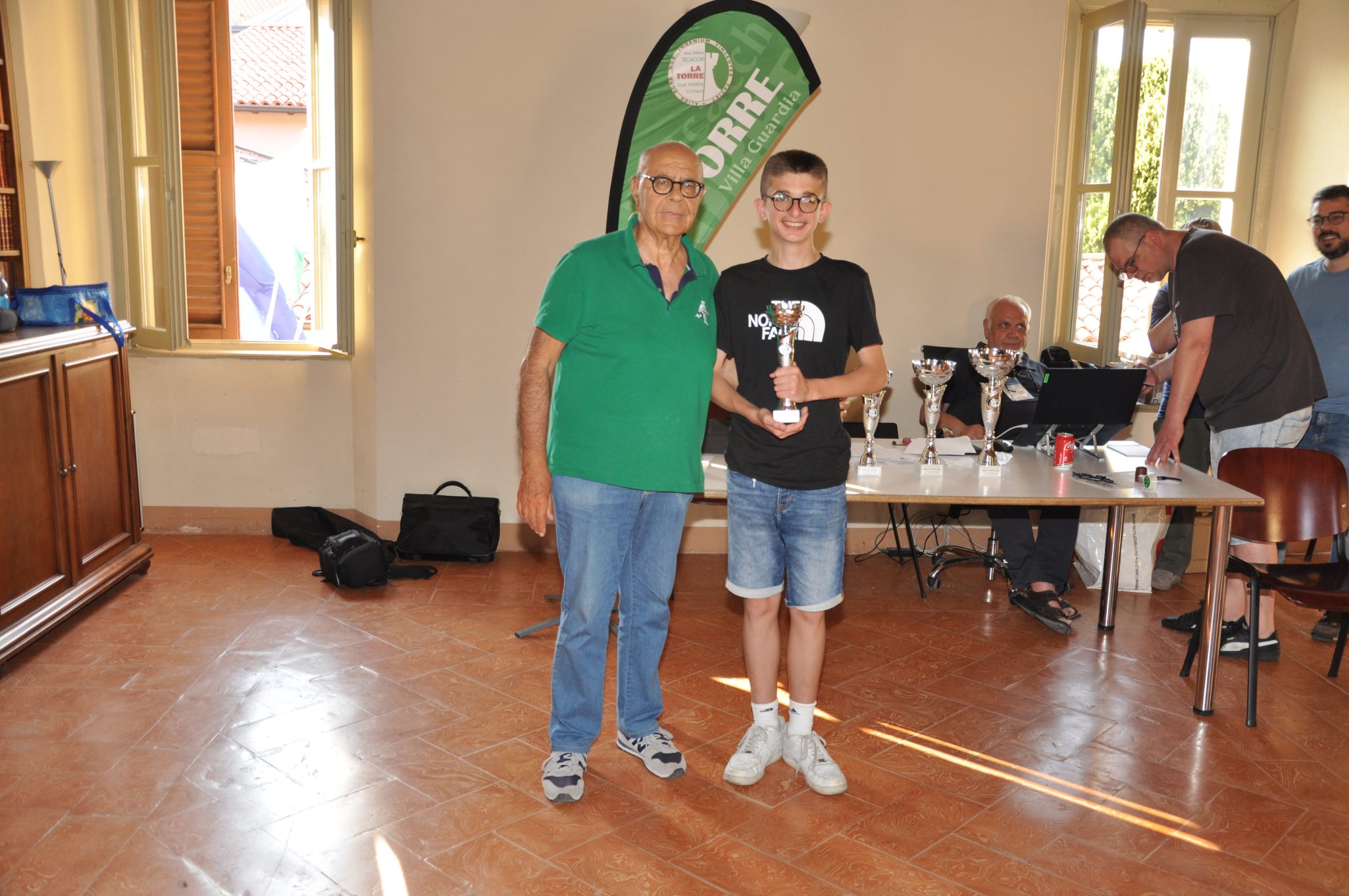 Un adulto in maglietta verde posa accanto a un giovane che tiene un trofeo, all'interno di una sala con finestre aperte e tavoli. Sullo sfondo, ci sono tazze e computer.
