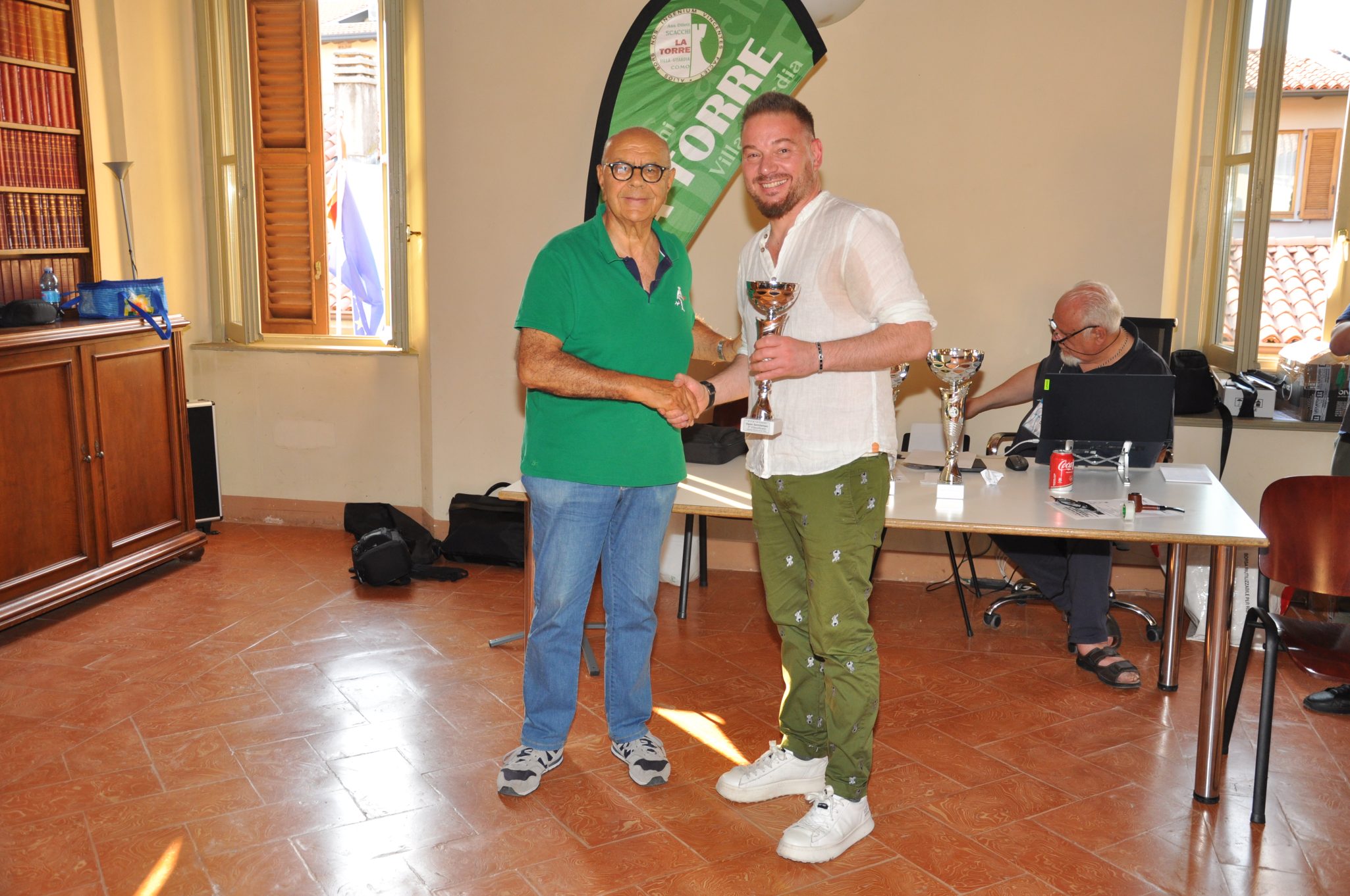 Due uomini si stringono la mano in una cerimonia di premiazione, uno dei quali tiene un trofeo. Sullo sfondo si vedono trofei e un computer su un tavolo.