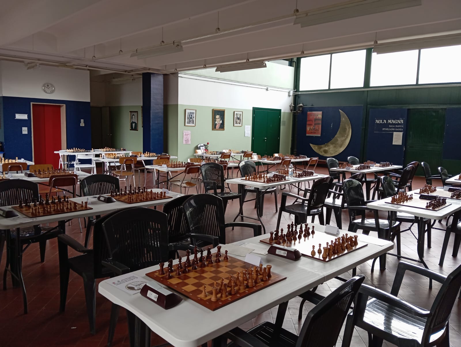 Interno di una sala con tavoli e sedie, pronti per un torneo di scacchi. Alcuni tavoli sono apparecchiati con scacchiere e pezzi in legno. Sullo sfondo si vedono pareti decorate e finestre che lasciano filtrare la luce.