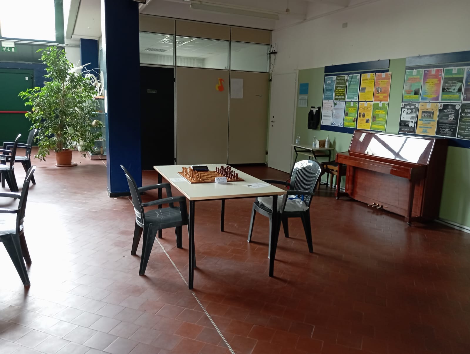 Un'area interna con tavolo da scacchi e sedie, una pianta in vaso e un pianoforte in fondo, circondata da poster sulle pareti.
