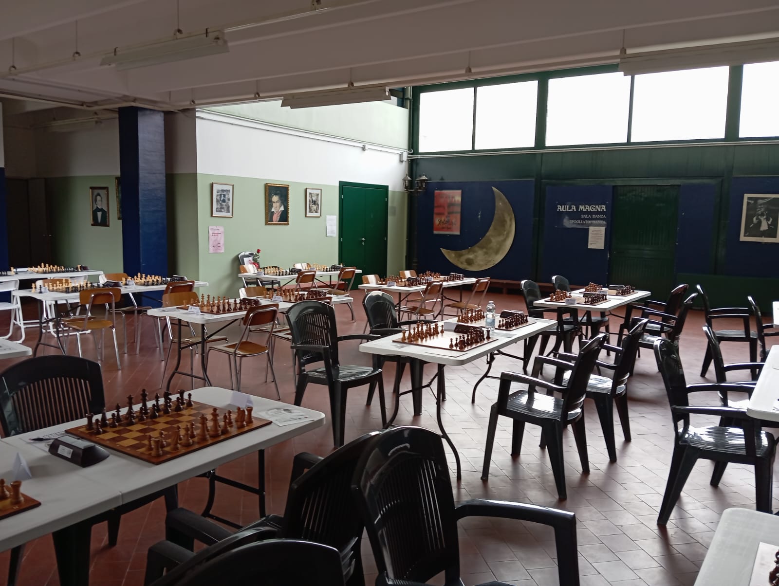 Interno di una sala da gioco a scacchi, con tavoli e scacchiere preparate. Sedie nere sono disposte intorno ai tavoli e ci sono ritratti alle pareti.