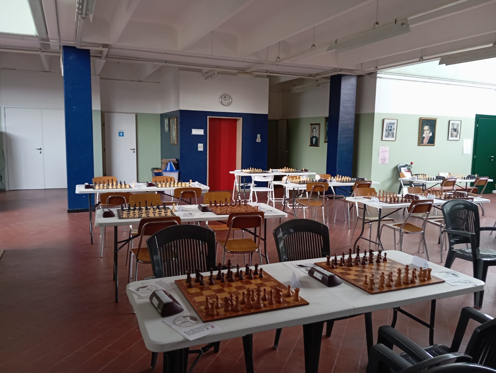 Una sala di gioco a scacchi con tavoli e scacchiere pronte per l'uso, sedie disposte attorno ai tavoli e pareti decorate con quadri.